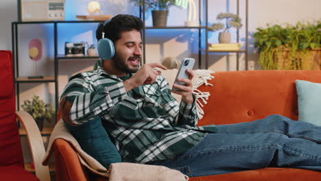 Happy-indian-man-in-wireless-headphones-relaxing-sitting-on-sofa-at-home-listening-favorite-music