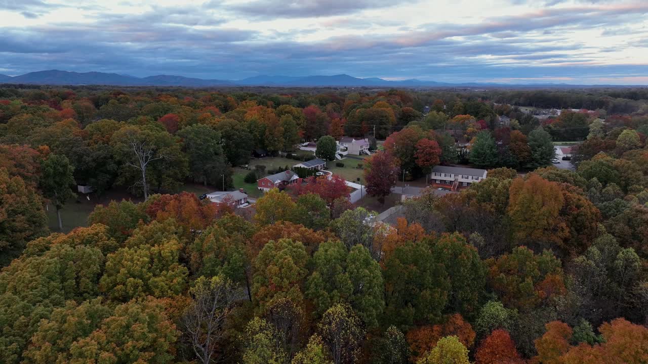 Aerial approaching shot of American community between colored trees in autumn. Cloudy dusk scene. Apartment and one family houses in American neighborhood. Wide shot