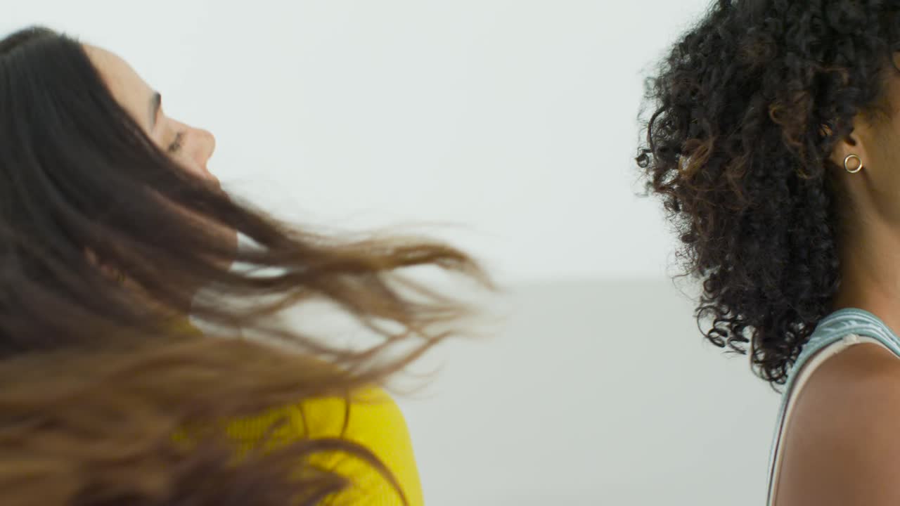Mulheres diversas ensaiando em estúdio, uma cacheada de listras levantando o braço, e a parceira de amarelo balançando o cabelo.
