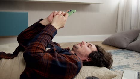un tipo con el cabello rizado en una camisa a cuadros se desplaza a través de las redes sociales mientras está tendido en el suelo en una almohada cerca de una computadora portátil y una taza de café en casa