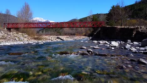 Volando-Río-Arriba-Y-Pasando-Por-Debajo-Del-Puente-Rojo-En-Los-Pirineos-Españoles,-Con-Las-Montañas-Nevadas-Al-Fondo