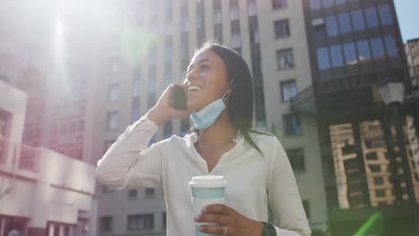 Mujer-Afroamericana-Con-Mascarilla-Hablando-En-Un-Teléfono-Inteligente-Sosteniendo-Café