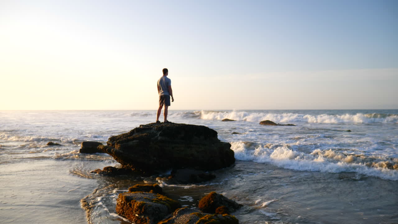A man watching the morning sunrise on a rock as ocean waves crash in slow motion on the beach in Santa Barbara, California