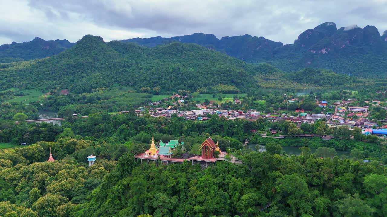 Drone orbits around a temple on a mountain summit outside Sangkhlaburi, Thailand, capturing views of the forest, river, town, and distant mountains