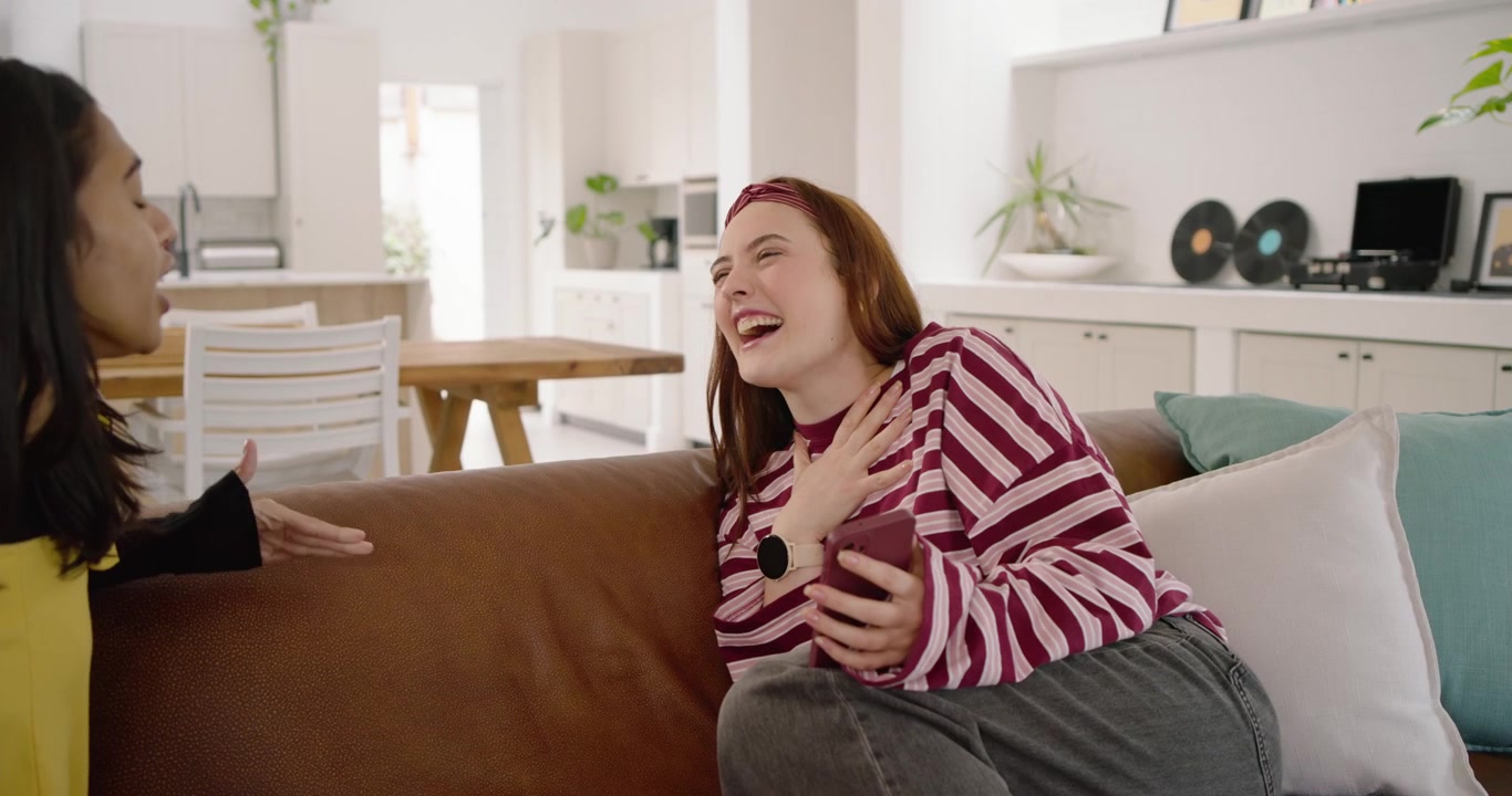 Diverse female friends on brown couch, right woman showing phone explaining while left leaning in