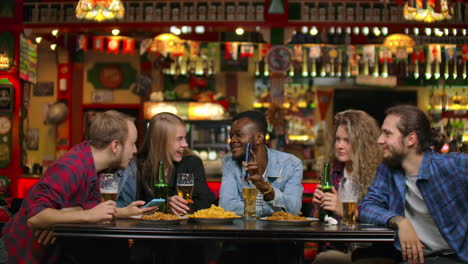 african american shows friends pictures on the phone they look and laugh while sitting in a bar in slow motion. company of young men and women.