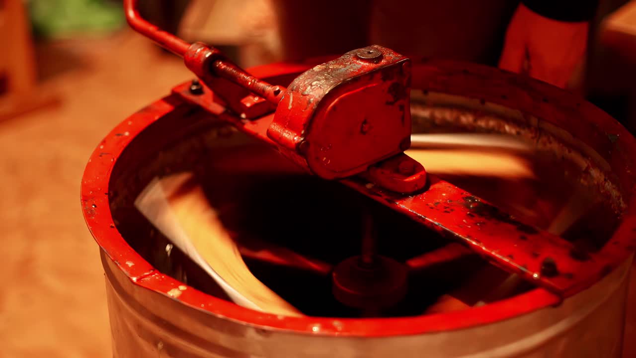 Static slow motion of hands turning a hand crank mixer in a red bucket, rotating paddles blending thick liquid paste in a workshop or small production setting