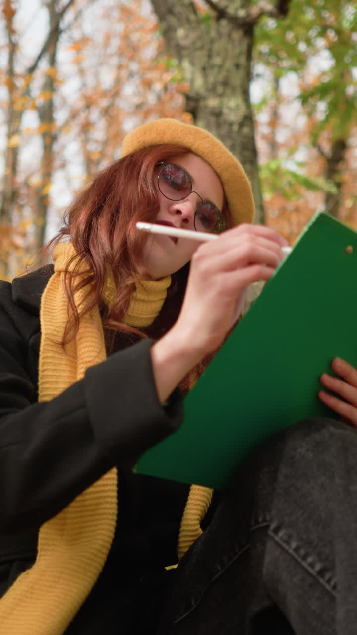 artista dibujando pensativamente en lienzo, sentado con la cabeza inclinada hacia la izquierda en un banco de parque de madera, rodeado de follaje de otoño, vestido con elegante boina amarilla y bufanda