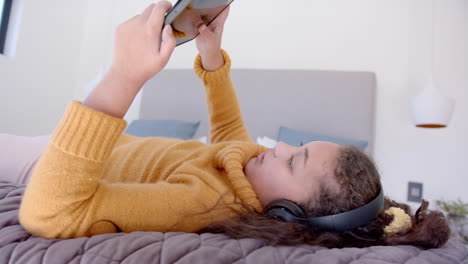 chica biracial feliz acostada en la cama, usando tableta y auriculares en un dormitorio soleado