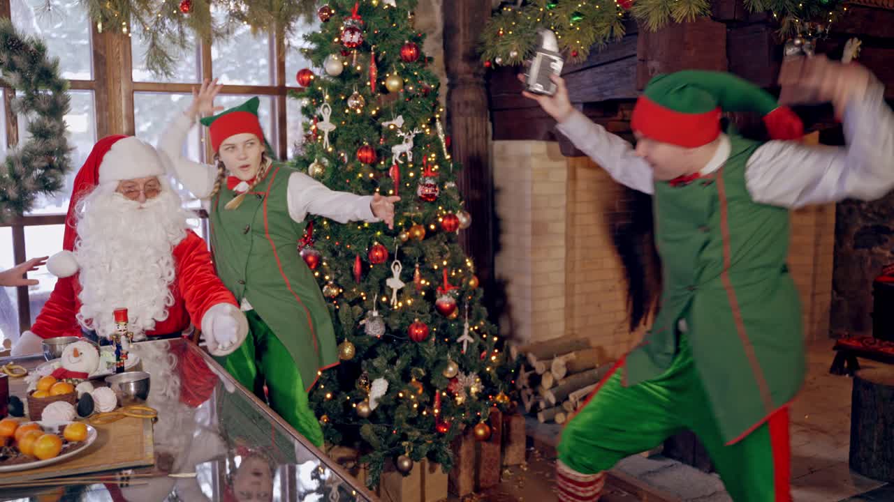 Happy elf and Santa near the Christmas tree. Elf is taking photo of a happy fairy characters in decorated room.