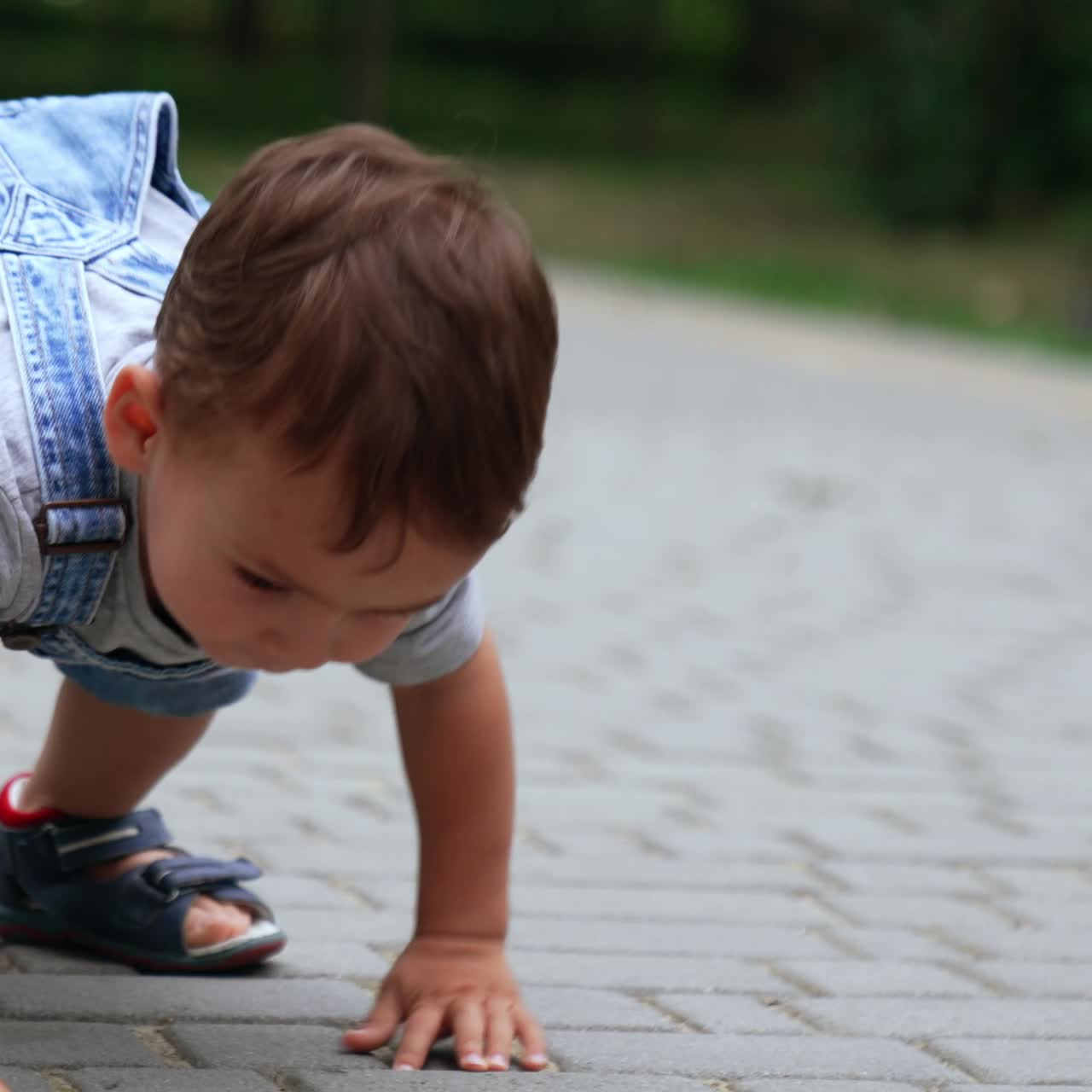 Lovely Caucasian boy sit squatted on the paved road. Adorable kid touches the tiles and crawls on his hands and feet