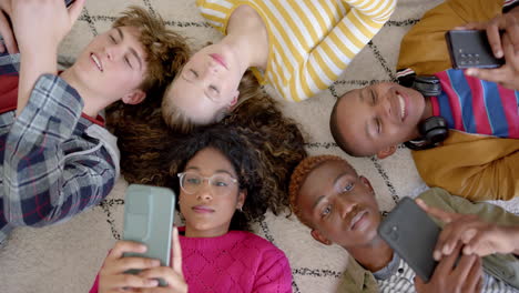 happy diverse group of teenage friends using smartphones and lying on carpet at home, slow motion