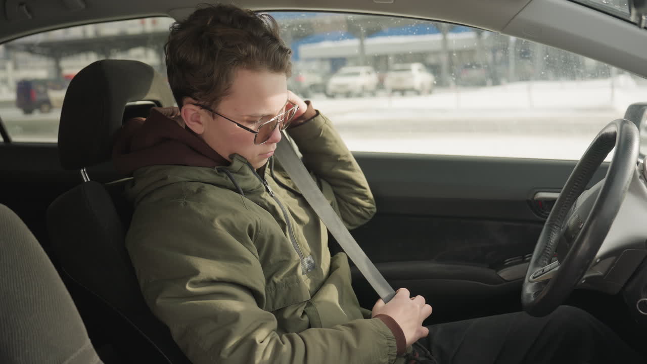 side view teenager putting on seat belt inside car with green jacket, winter light illuminating interior, building and parked cars visible through window, capturing safety routine