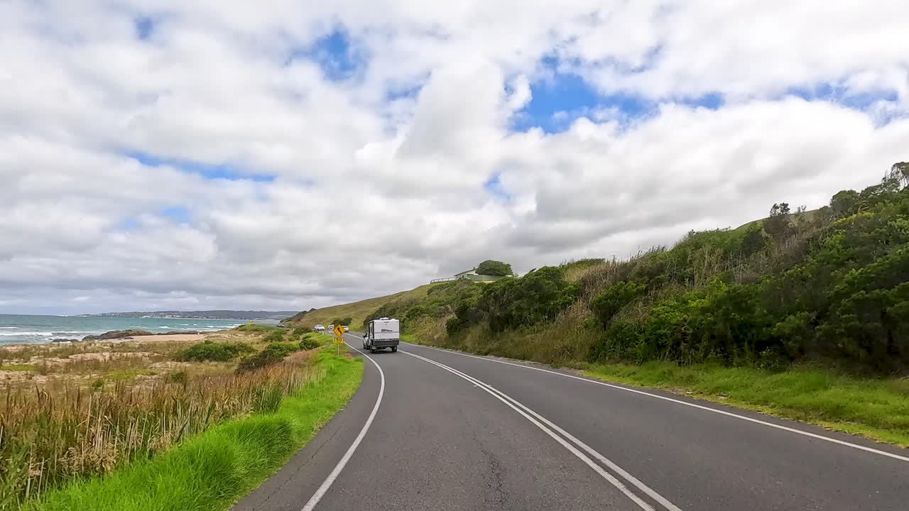 A vehicle travels the picturesque Great Ocean Road, showcasing coastal views and lush greenery under a partly cloudy sky