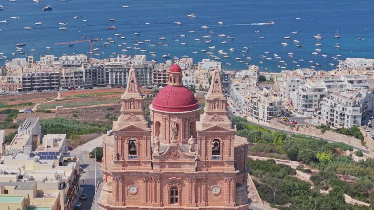 An aerial view of the Mellieha Parish Church in Malta, featuring its red dome and twin bell towers. The church overlooks a vibrant coastal town with numerous boats anchored in the deep blue waters