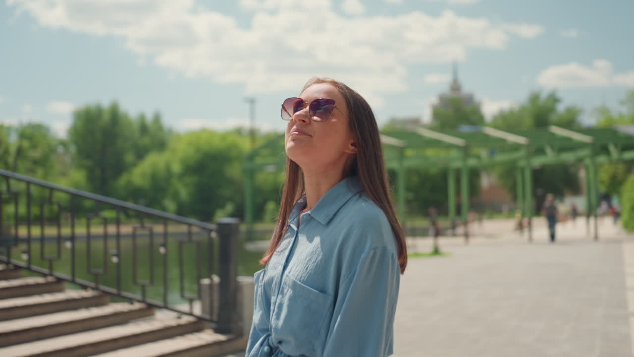 Mujer mirando al cielo alegremente en el exterior, mujer caucásica radiante sonriendo al cielo durante un paseo animado, mujer de piel clara con gafas de sol sonriendo hacia arriba en medio de un alegre descubrimiento al aire libre y un clima soleado