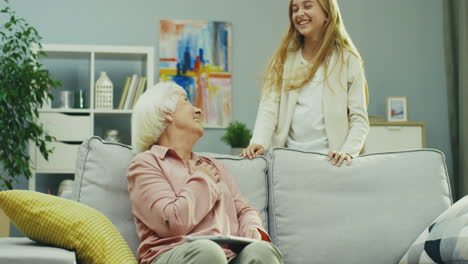 portrait shot of the old good looking woman sitting on the sofa and holding tablet in the room at home while her cute teenager coming to her from behind, leaning on the couch and they smiling