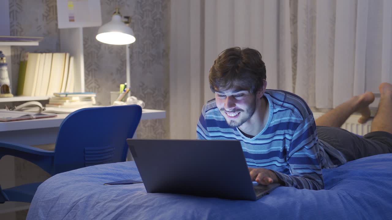 un joven tendido en la cama mirando una computadora portátil.