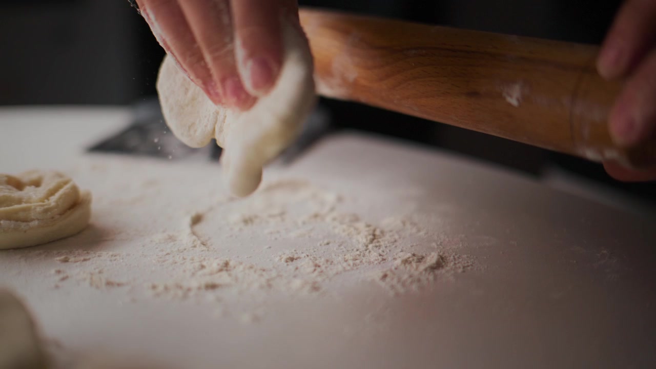 Mani stendono l'impasto con un mattarello di legno su una superficie infarinata. Le dita guidano l'impasto mentre si appiattisce. La scena cattura un momento accogliente e concentrato in cucina. Perfetto per video di ricette, tutorial di cucina o contenuti di pasticceria