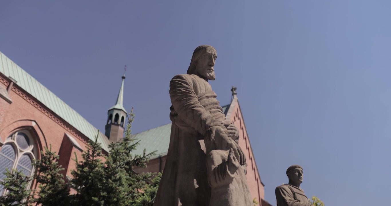 Un'inquadratura dinamica di una statua di marmo di un santo apostolo che tiene la mano di un bambino, con una chiesa e alberi sullo sfondo in una soleggiata giornata estiva, risoluzione 4K e slow motion.
