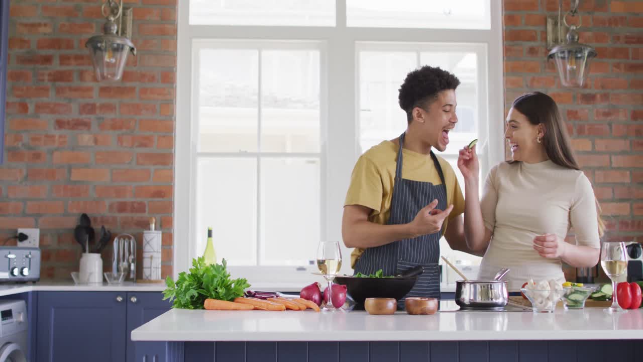 feliz pareja biracial cocinando juntos y riendo en la cocina