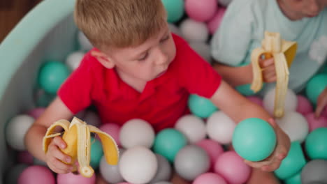 overhead view of little kids sitting in colorful ball pit eating bananas, boy in red shirt and girl in pastel clothes enjoying playful snack time with bright light