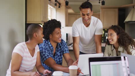 group of diverse male and female friends in kitchen, laughing and working on laptops