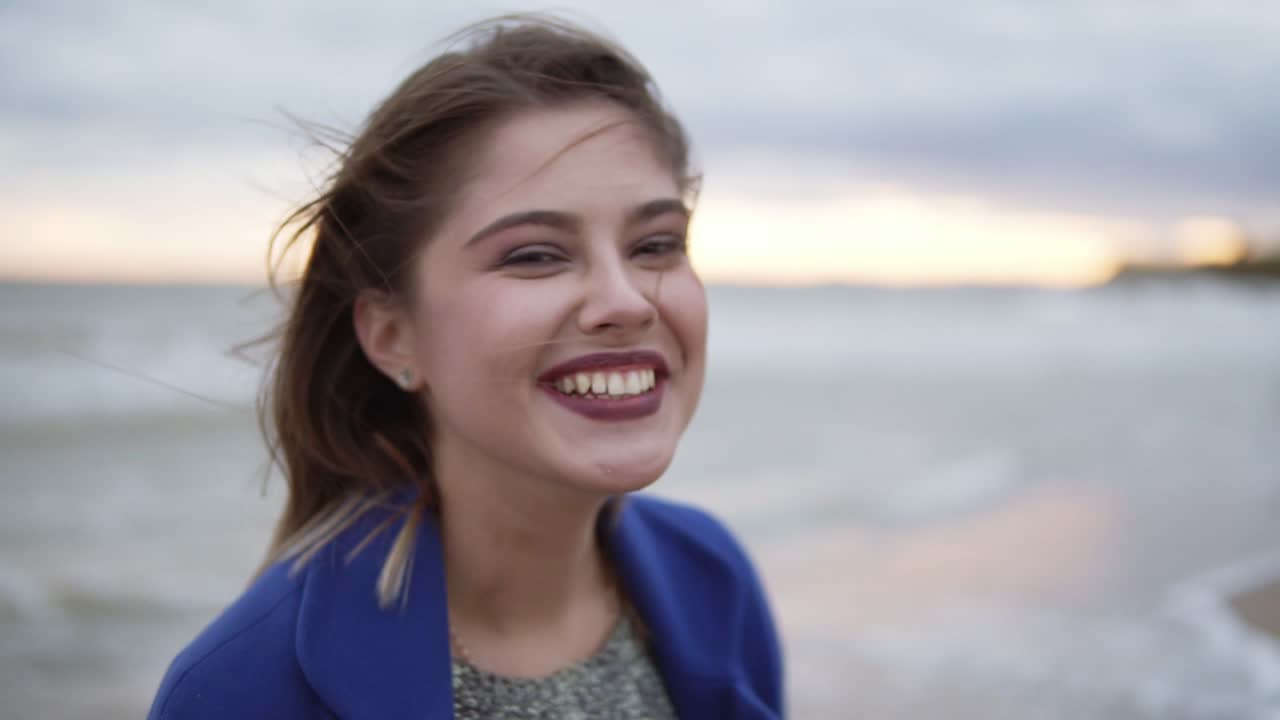 mujer joven sonriente atractiva con hermoso maquillaje de pie en el viento contra el paisaje del mar y mirando a la cámara. cámara lenta