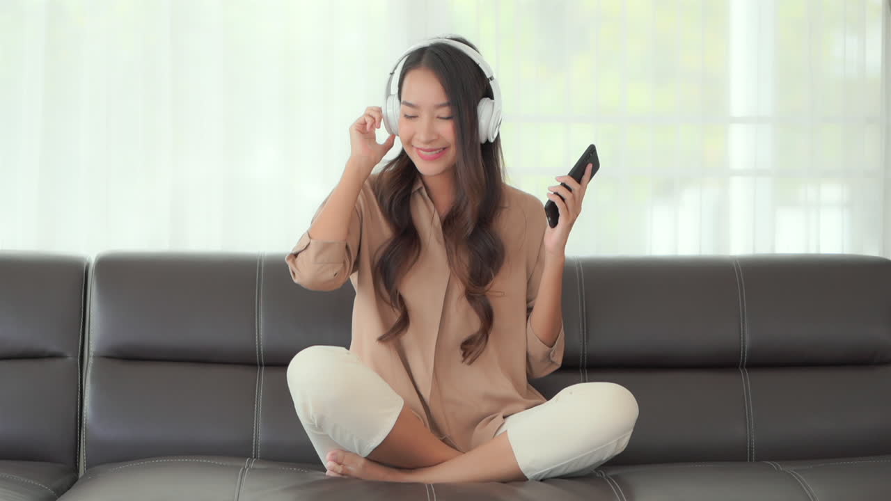 A lovely Asian girl wearing on-ear headphones is listening to music and dancing while holding her cellphone in hand, excited. Sitting on the sofa with her legs crossed