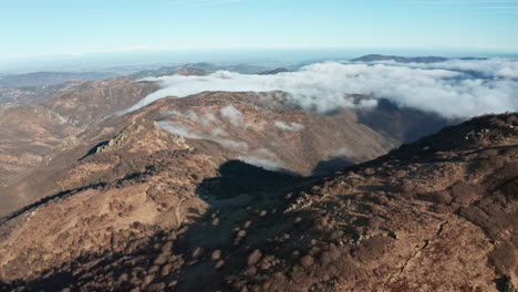 Mountain-landscape-with-clouds-rolling-over-peaks,-casting-shadows,-clear-skies-above,-aerial-perspective