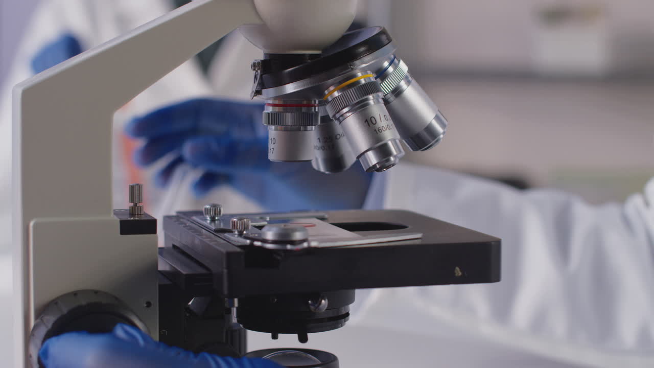 Close Up Of Female Lab Worker Or Phlebotomist Analysing Blood Samples In Laboratory With Microscope