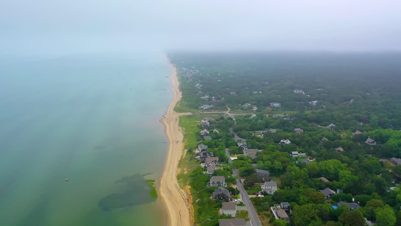 Vista aerea panoramica della spiaggia di Cape Cod sotto nuvole pesanti e nebbia leggera, con costa sabbiosa che si estende verso l'orizzonte, onde oceaniche calme e quartieri residenziali lungo la costa