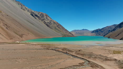 Drone footage captures a sweeping mountain landscape with a vibrant turquoise lake and rugged slopes under a vivid blue sky on a calm, while a vehicle travels a solitary journey