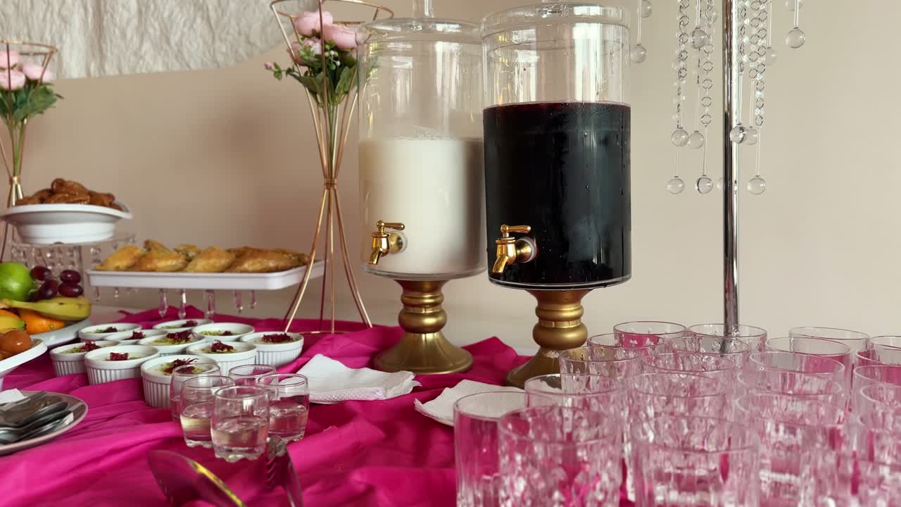 Dispensadores de bebidas de cristal llenos de zumo oscuro y leche expuestos en una mesa de bufé de catering decorada con pasteles, postres y copas de cristal bajo una iluminación interior suave