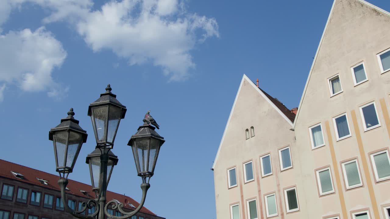 Camera tilts upward revealing ornate street lamps, angular buildings, blue sky, and scattered clouds