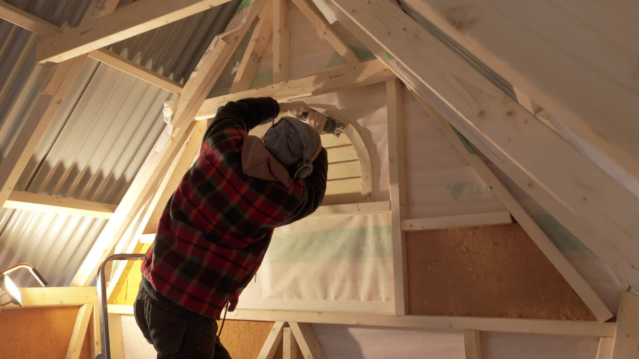 Handyman med hørselvern bruker stikksag for å kutte et halvmåneformet vindu i et lite trehus.