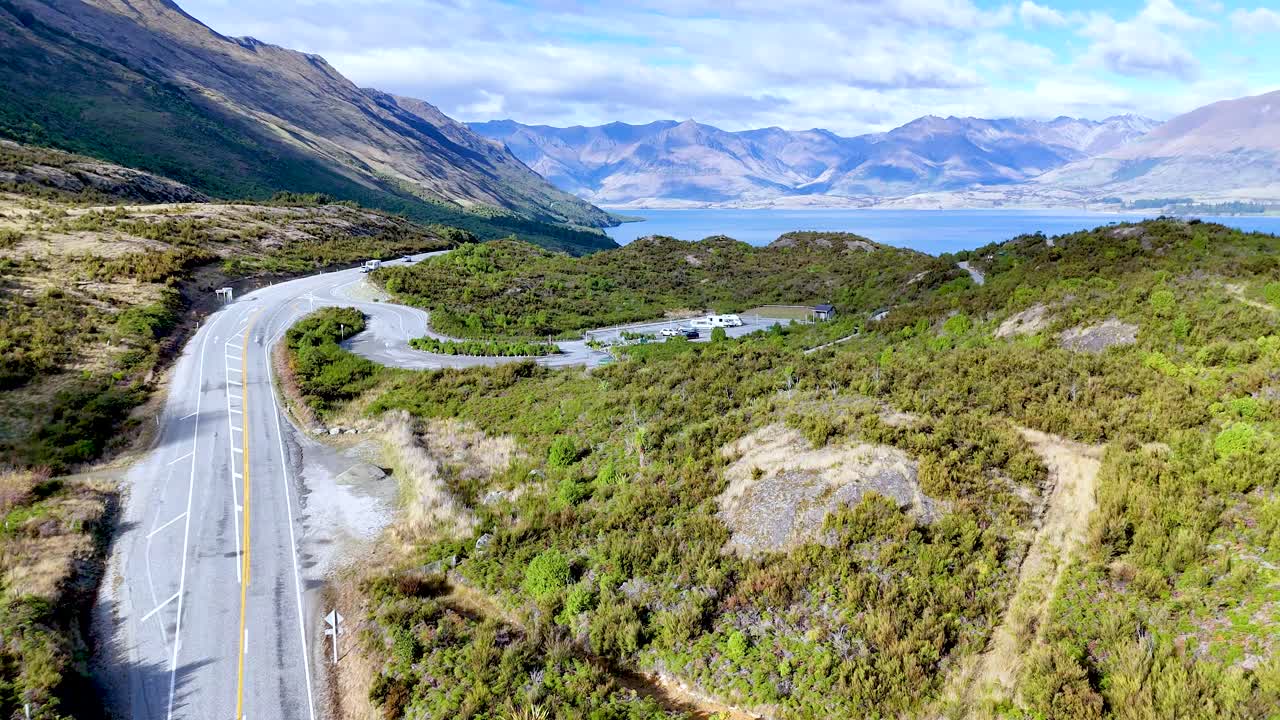Drone glides above curving mountain road, lush greenery, and lake under bright daylight