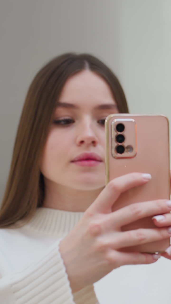 Signora con maglione bianco che scatta una foto con lo smartphone, espressione concentrata, interni moderni sullo sfondo con illuminazione soffusa e design elegante, ambiente minimalista con piante decorative verdi