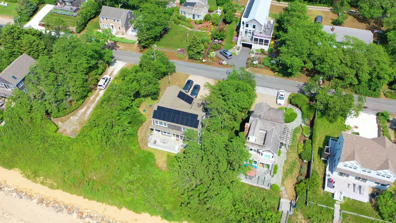 Drone reveals Cape Cod summer life with a beachfront property overlooking golden sand, families below, and turquoise water glimmering beside lush greenery