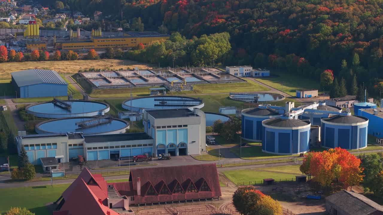 Aerial shot of a wastewater treatment plant in Kaunas, Lithuania, surrounded by colorful autumn forest, showcasing circular tanks and modern water purification infrastructure
