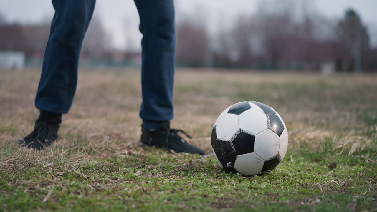 Close-up leg view of two individuals engaged in a soccer game as one person kicks the ball to the left, with a blurred background of grassy field
