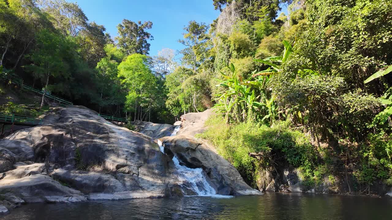 Svelamento graduale della Cascata Mor Paeng a Pai, Thailandia. Una pozza d'acqua serena riflette la luce solare su rocce scoscese circondata da vegetazione lussureggiante. Cattura di una bellezza naturale pacifica e incontaminata.