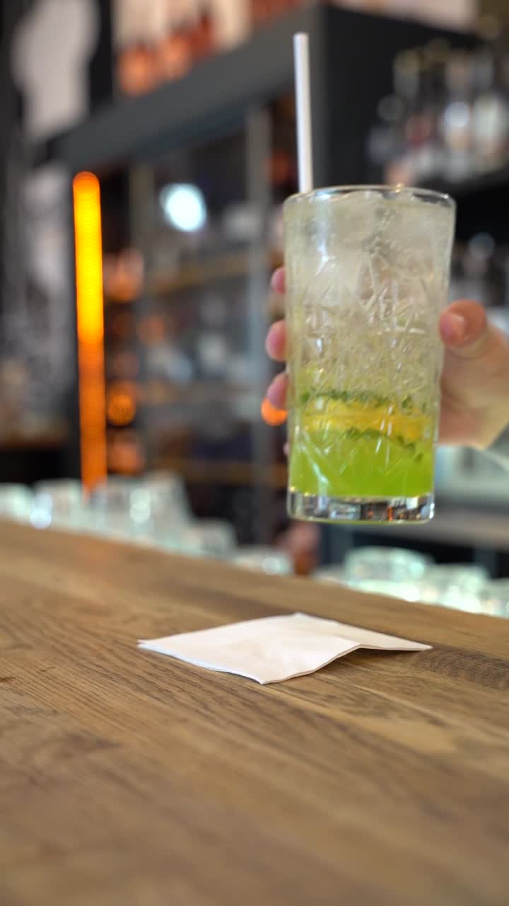 A vibrant lime soda cocktail served in a textured glass sits on a wooden bar counter. The drink is garnished with fresh lime slices and ice, with a straw ready for sipping.