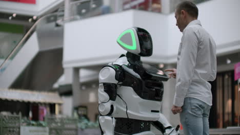 un hombre se para con un robot bot y le hace preguntas y pide ayuda haciendo clic en la pantalla en el cuerpo del robot. la interacción humano-robot en el mundo moderno