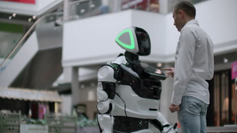 a man stands with a robot bot and asks him questions and asks for help by clicking on the screen on the robot body. human-robot interaction in the modern world