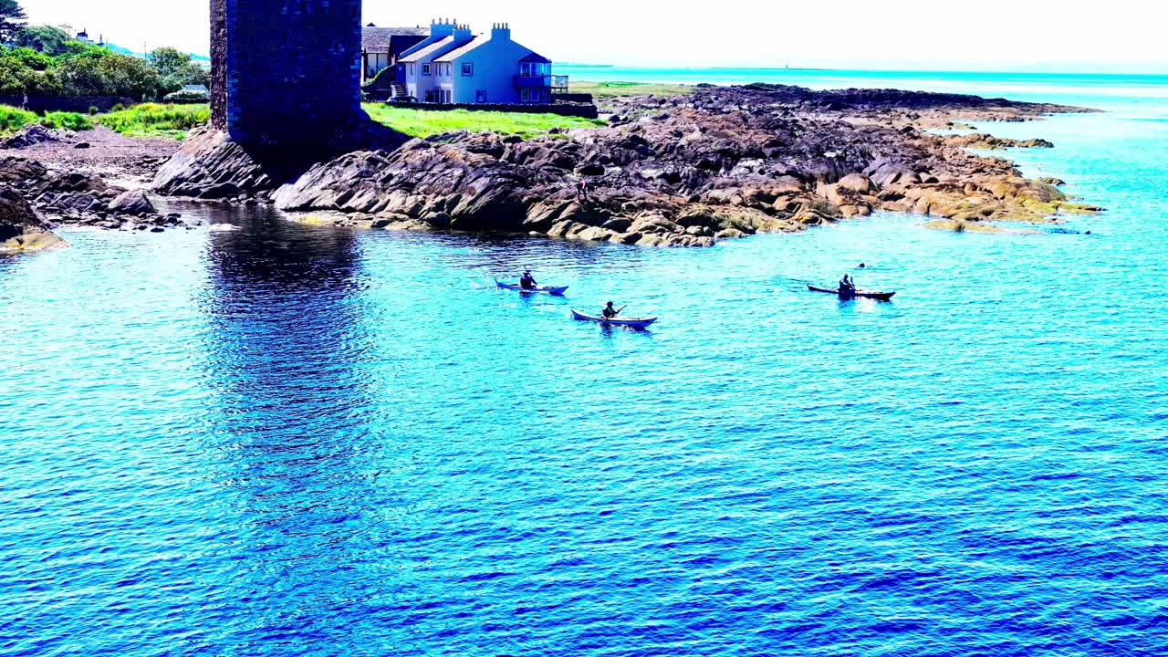 Filmato aereo di kayakisti che scivolano attraverso acque costiere limpide accanto a una costa rocciosa e al Castello di Portencross vicino a Largs, Scozia, catturando il paesaggio marino estivo e l'avventura all'aria aperta.
