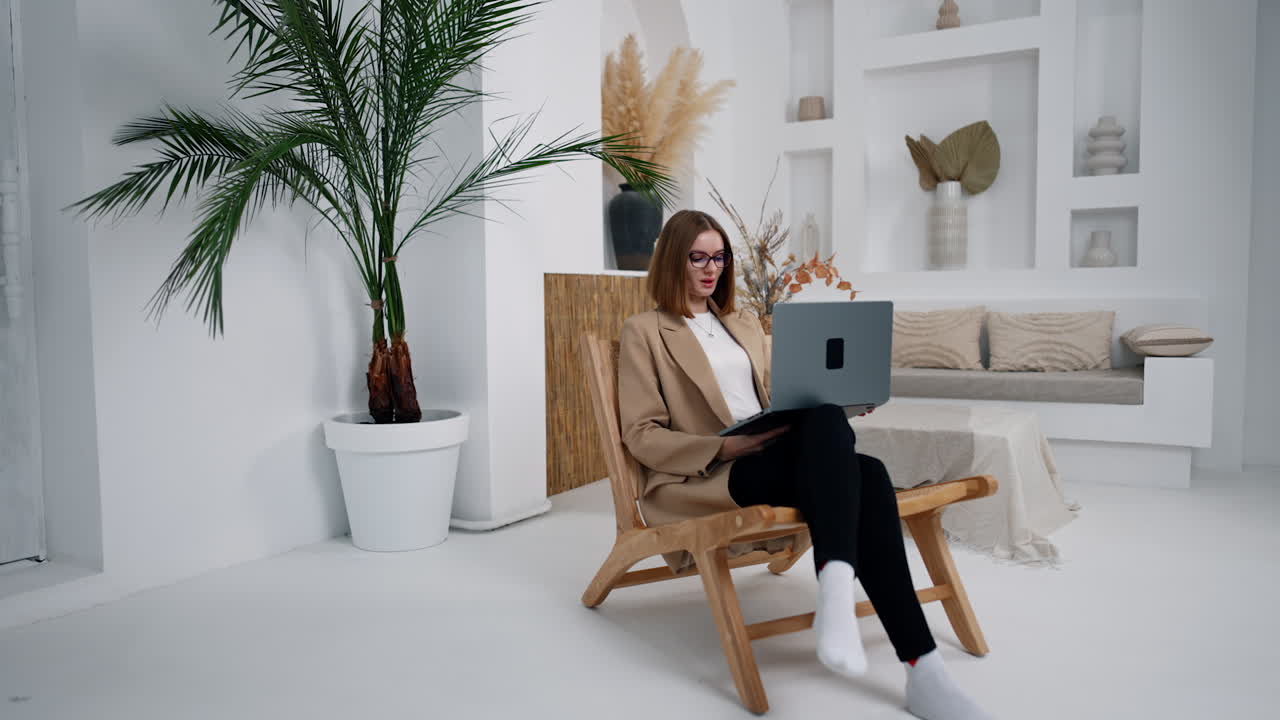 Businesswoman sitting in the armchair with laptop in hands. Modern woman working in comfortable light room.