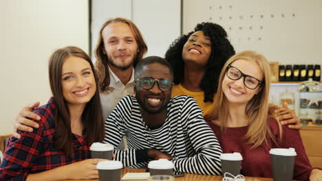 grupo multiétnico de amigos sonriendo y mirando la cámara sentados en una mesa en un café