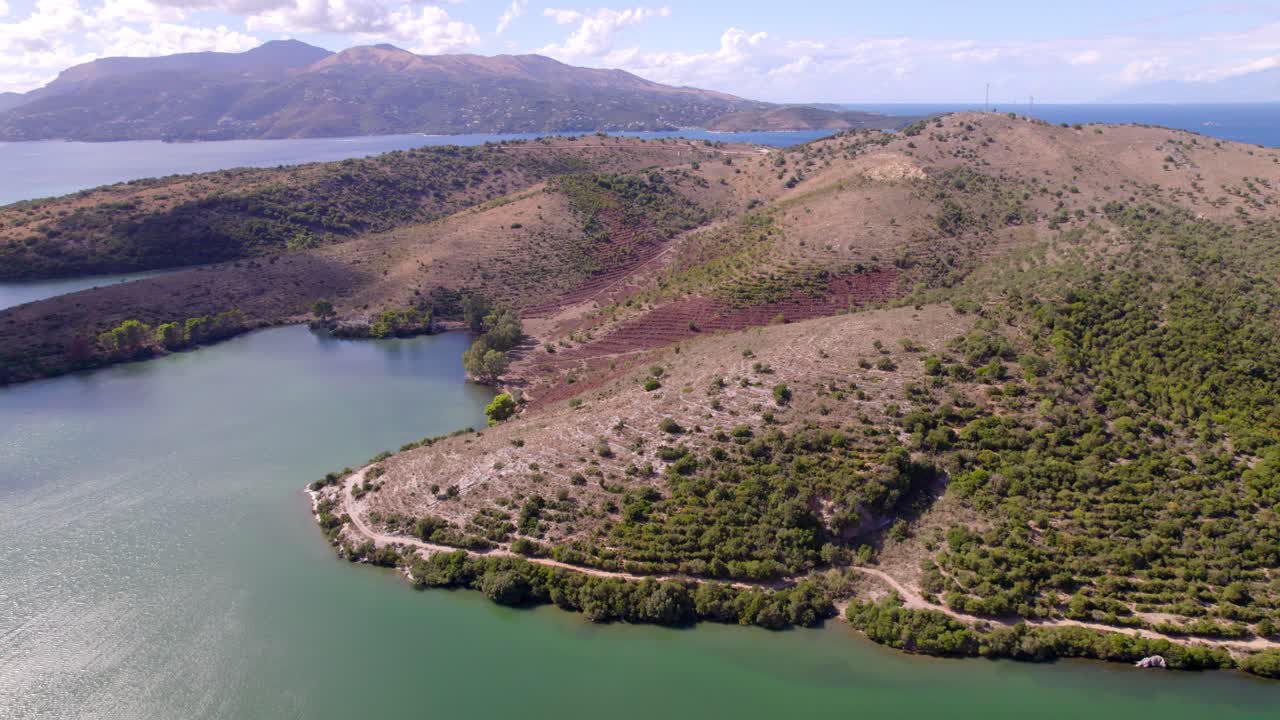 Vídeo de dron muestra una península escarpada con colinas cubiertas de maleza junto a una laguna tranquila y mar abierto en la distancia bajo un cielo parcialmente nublado, movimiento suave hacia adelante del dron