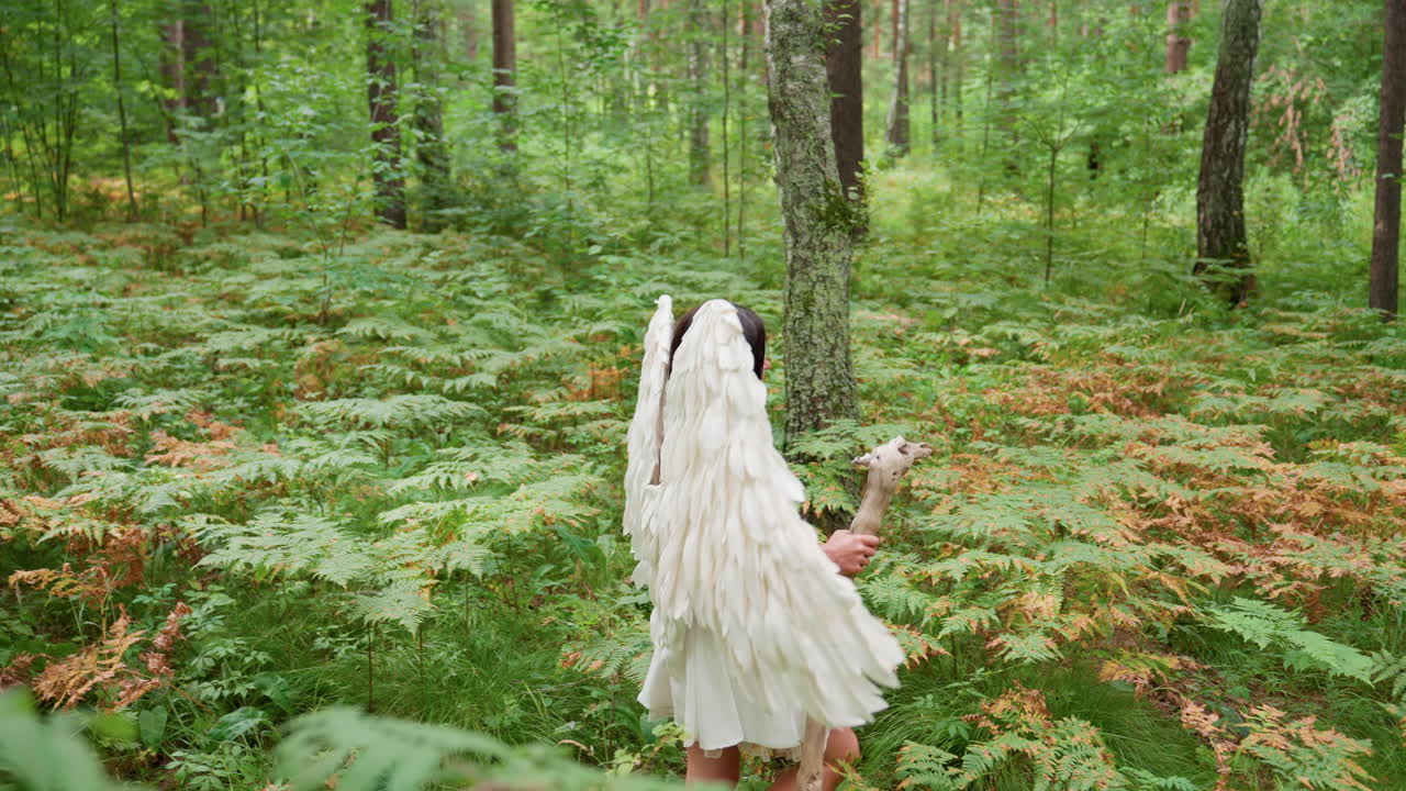 Female forest guardian dressed in white with large angelic wings walks peacefully along green fern covered forest path holding wooden staff, exuding calm and mystical grace in magical woodland